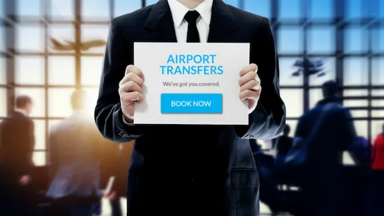 Hombre de negocios sosteniendo un cartel de "Airport Transfers" en una terminal de aeropuerto. Servicios de traslados al aeropuerto, transporte privado y reservas online.